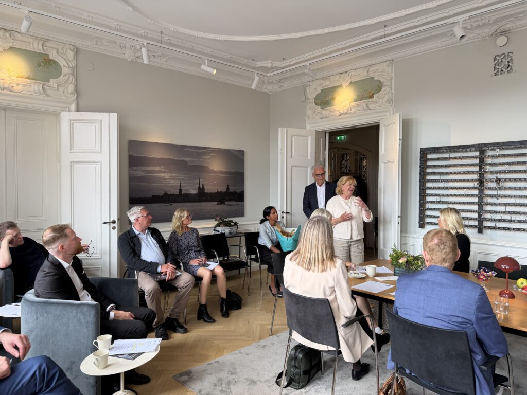 Deltagare samlade i ett klassiskt rum på Munkbron under Änke- och Pupillkassans årsstämma. En kvinna talar stående inför gruppen, med en stor Stockholmsfotografi på väggen bakom.