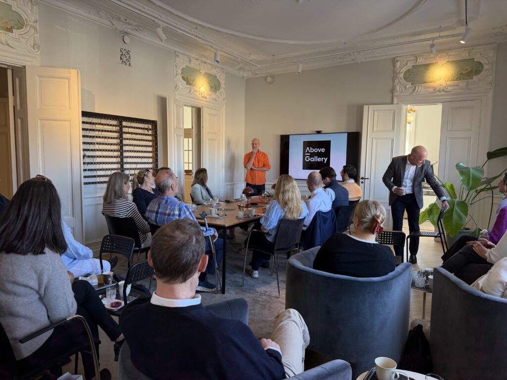 Tema konst med Anders Kumlien från Above Gallery presenterar för en grupp deltagare under Frukostklubben hos Änkan. En person i orange tröja står framför en skärm med Above Gallery-logotypen.
